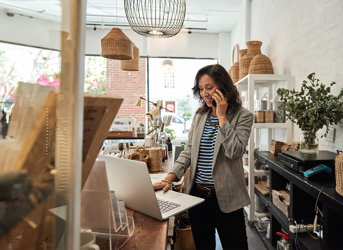 Business Insurance - Portrait of a Mature Business Woman Standing in Her Main Street Shop While Using a Laptop and Talking on the Phone